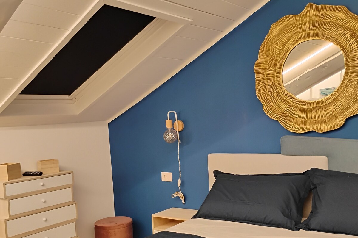 A bedroom with a sloped ceiling, a bed with dark bedding, a wooden dresser, a round mirror, and a wall-mounted lamp.