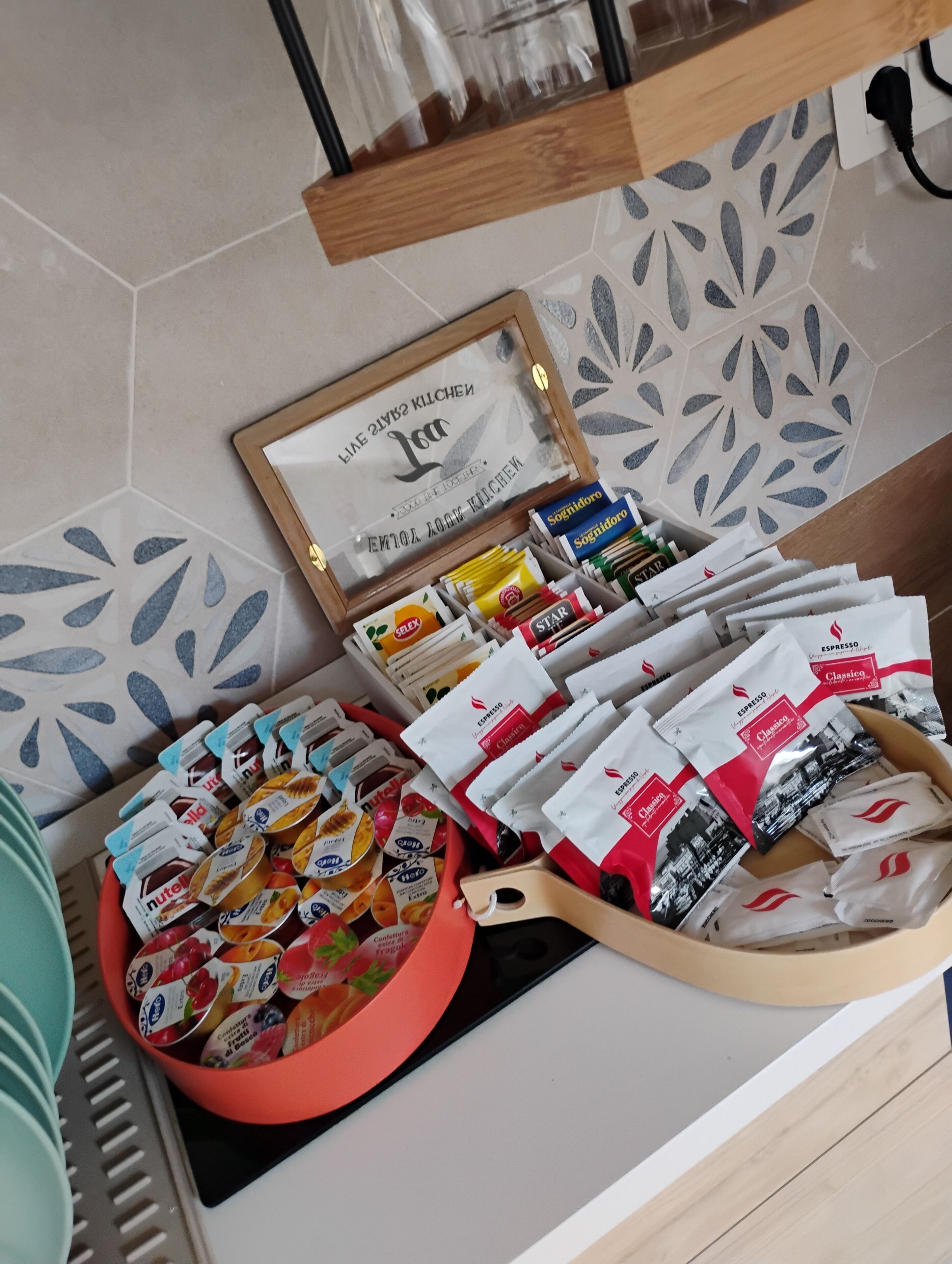 Een aanrecht met verschillende verpakte levensmiddelen, een lijst in een fotolijstje, en een decoratieve tegelachtergrond.