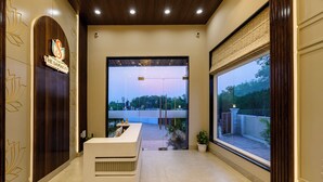A reception area with a reception desk, a large glass door leading outside, and a view of the outside. - Amritara The Samagam (Pindra)