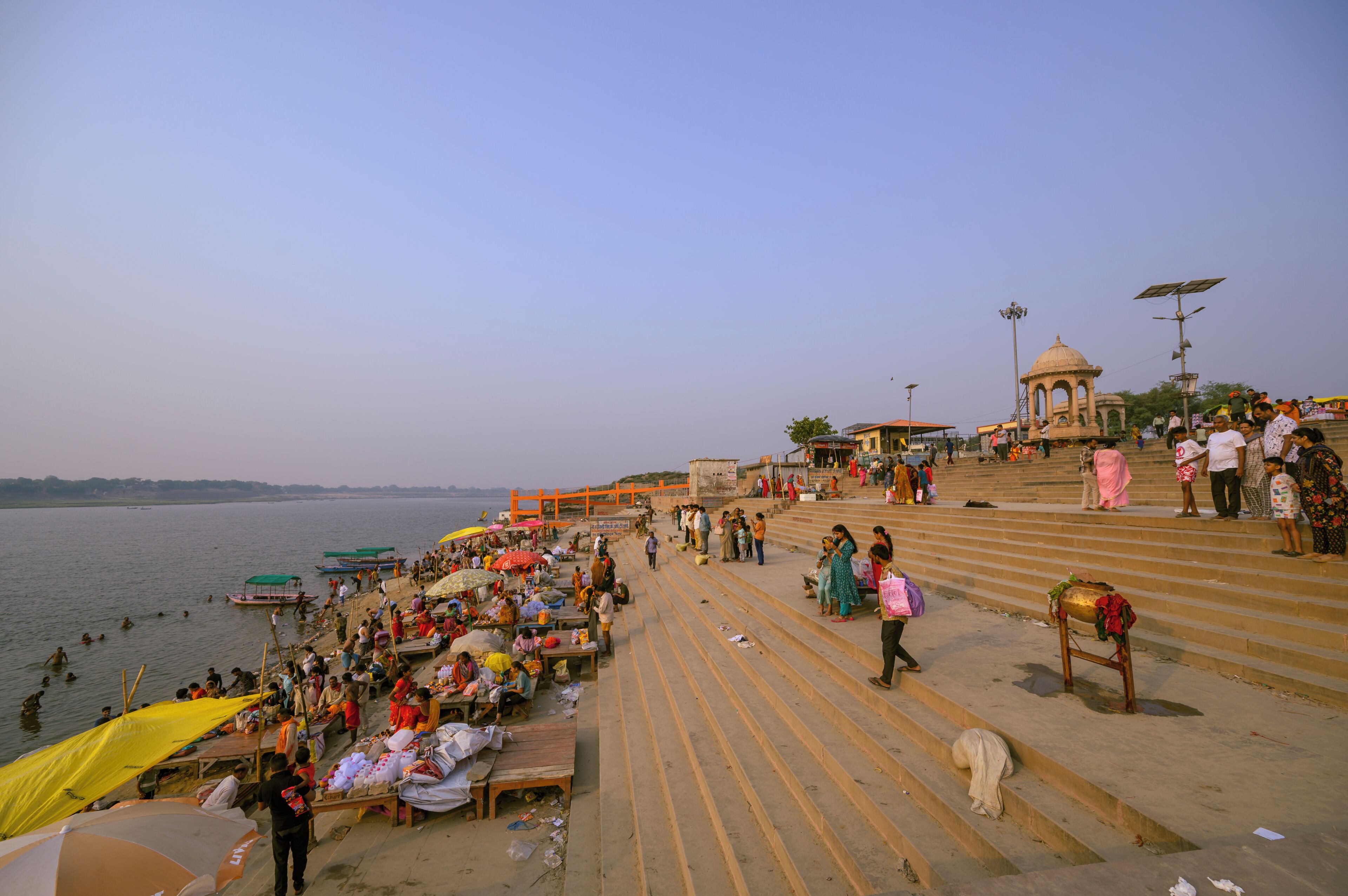 A riverside walkway with steps leading down to the water, a small pavilion, and people walking and sitting along the path.