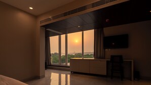 A hotel room with a large window, a flat-screen TV, a desk, and a chair. - Amritara The Samagam (Pindra)