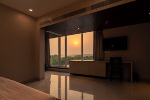 A hotel room with a large window, a flat-screen TV, a desk, and a chair. - Amritara The Samagam (Pindra)