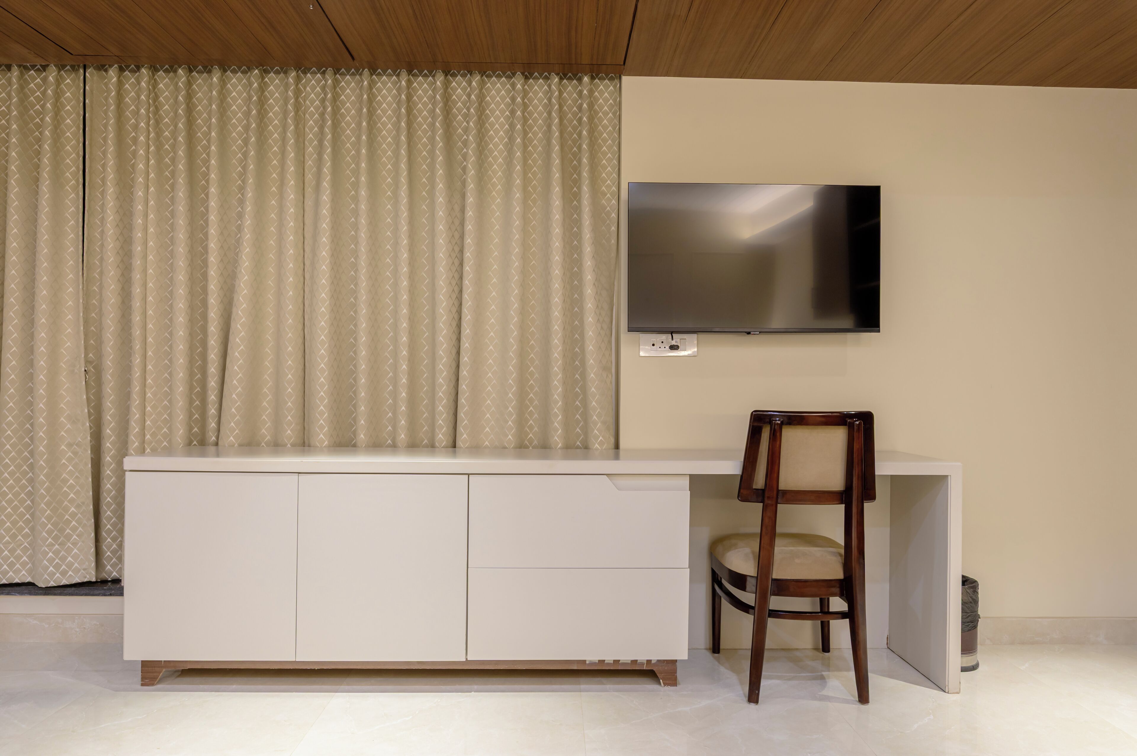 A room with a white cabinet, a TV mounted on the wall, a wooden chair, and a curtain.