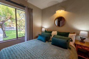 A bedroom with a bed, a nightstand, a lamp, a mirror, and a window with curtains. - Parsons River Camp (Hoedspruit)