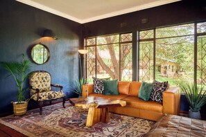 A room with a dark blue wall, a round mirror, a wooden coffee table, a leather sofa with green cushions, and large windows with a view of greenery. - Parsons River Camp (Hoedspruit)