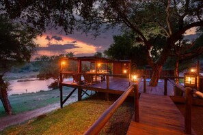 A wooden deck with a dining area and a view of a lake at dusk. - Parsons River Camp (Hoedspruit)