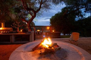 A stone fire pit with logs, a barbecue grill, and a wooden table with chairs in a wooded outdoor setting. - Parsons River Camp (Hoedspruit)