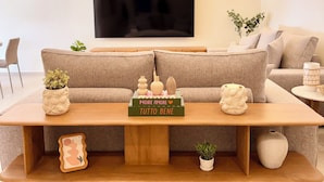 Uma sala de estar moderna com uma televisão de tela plana grande, uma mesa de centro de madeira com objetos decorativos e um sofá bege.