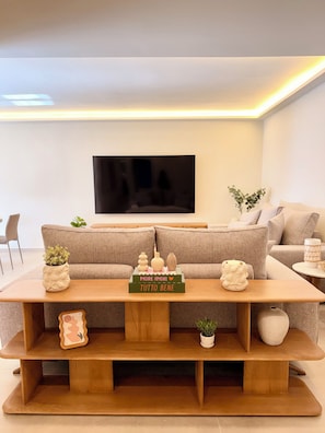 Ein modernes Wohnzimmer mit einem großen Flachbildfernseher, einem Couchtisch aus Holz mit Dekoration und einem beigefarbenen Sofa.
