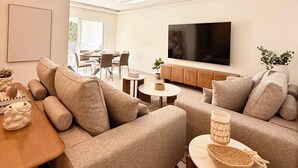 Uma sala de estar moderna com um sofá, uma mesa de centro, uma televisão e uma área de jantar ao fundo.