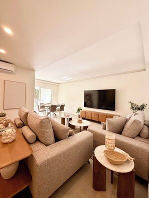 Ein modernes Wohnzimmer mit einer Sofagarnitur, einem Couchtisch, einem Fernseher und einem Essbereich im Hintergrund.