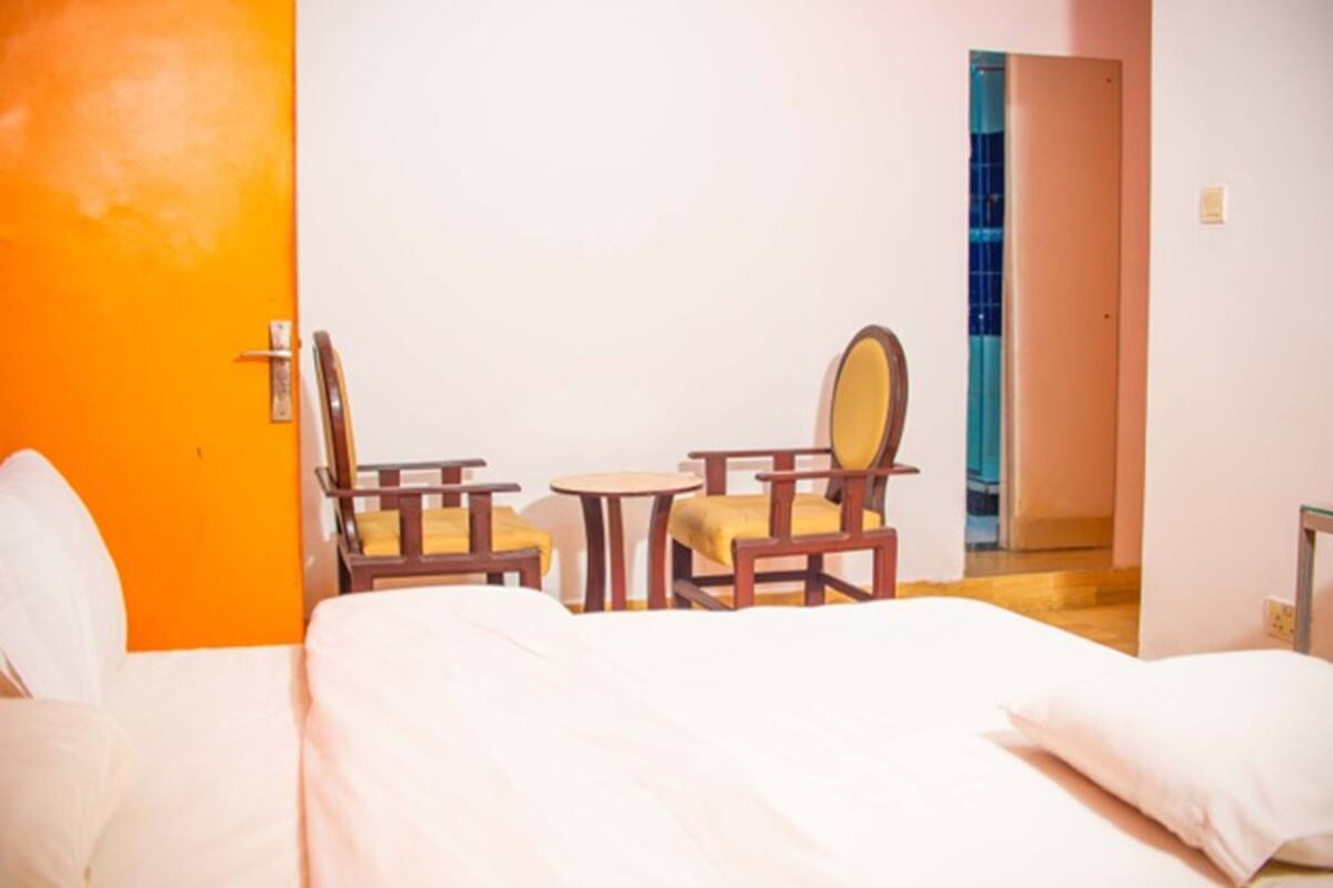 Habitación de hotel con cama, dos sillas de madera, una mesita redonda y una puerta que da a otra habitación.