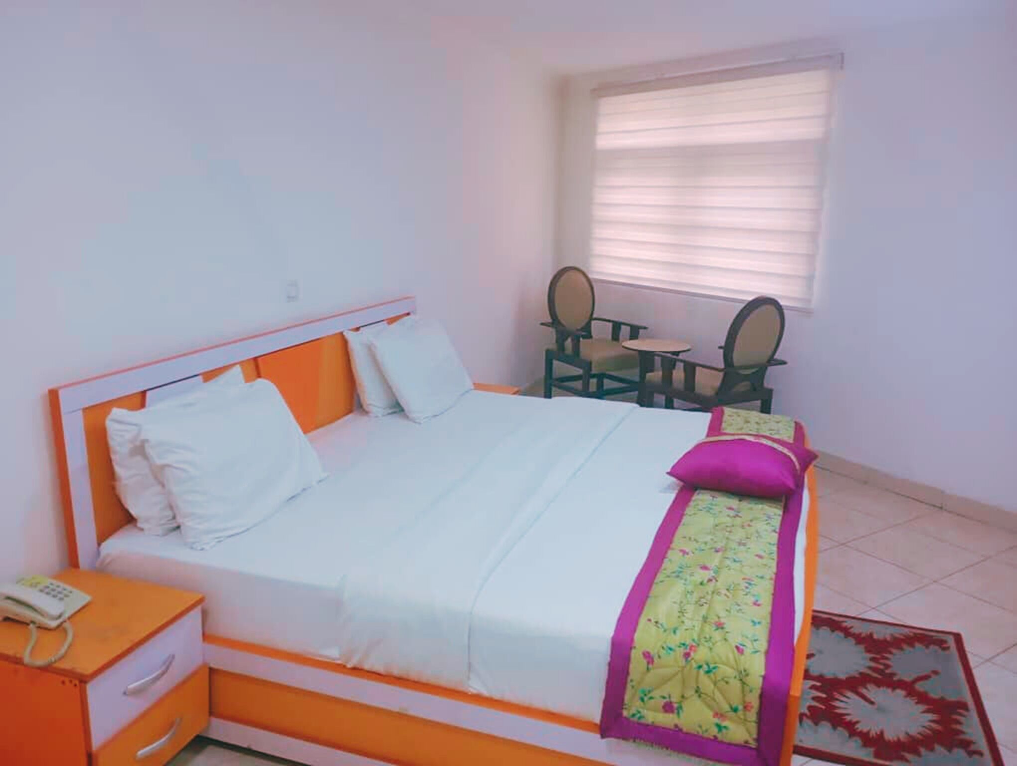 A bed with a white and orange headboard, a bedside table with a phone, and a window with blinds.