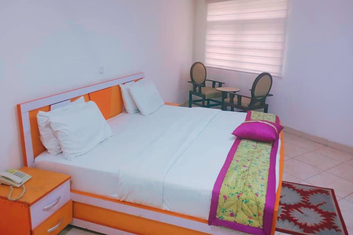 Una cama con cabecera blanca y naranja, una mesita de noche con un teléfono, y una ventana con persianas.