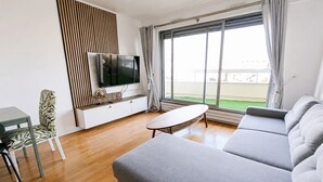 Ett modernt vardagsrum med en stor grå soffa, ett litet vitt bord, en tv på ett vitt skåp och utsikt över ett grönområde genom stora fönster.