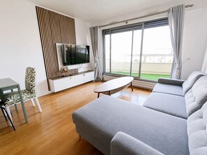 Uma sala de estar moderna com um sofá cinzento grande, uma mesinha branca, uma TV sobreposta a um armário branco, e vista para uma área externa verdejante através de grandes janelas.