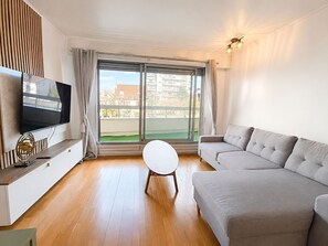Un salon moderne doté d’un téléviseur à écran plat, d’un canapé gris, d’une table basse ronde et d’une grande fenêtre offrant une vue sur les immeubles.