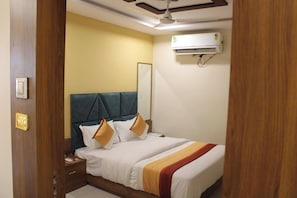 A hotel room with a bed, bedside table, nightstand, wall-mounted air conditioner, and a door with a number.