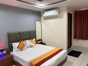 Ett hotellrum med en säng, ett sängbord, en liten avlastningsyta, en luftkonditioneringsanläggning, en spegel och ett fönster med gardiner.