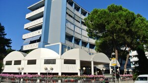 A multi-story hotel with balconies, a covered entrance, and a parking area. - Lovely Flat Just a Short Walk to the Beach (Lignano Sabbiadoro)