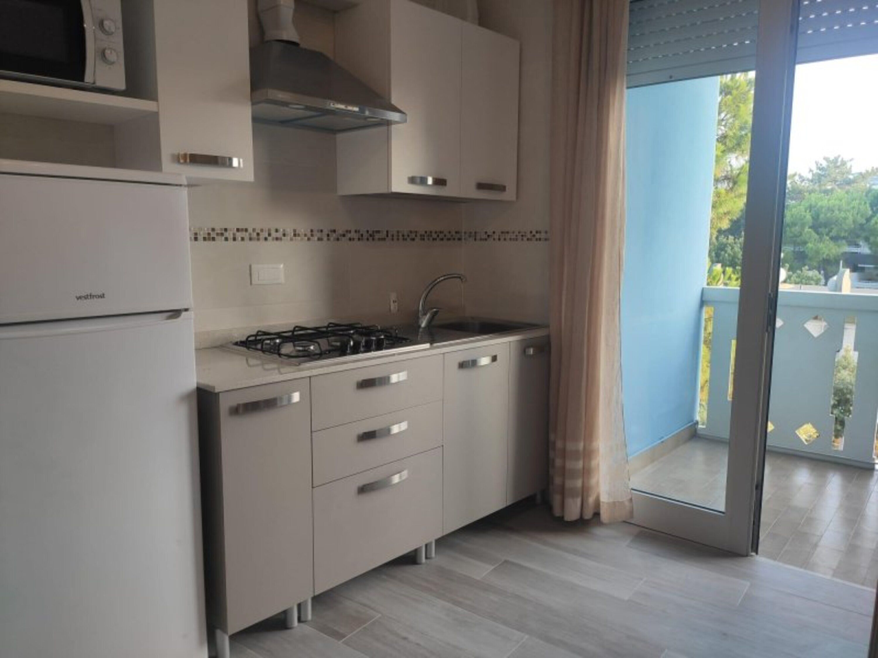 A compact kitchen with a refrigerator, stove, and sink, a window with a view of greenery, and a sliding glass door to an outdoor area.