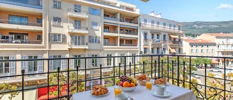 Un balcon avec une table dressée pour le petit-déjeuner, donnant sur une rangée d’immeubles à plusieurs étages.