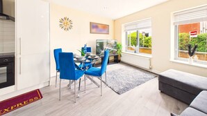 Een moderne keuken met witte kasten, een eethoek met blauwe stoelen, een televisie en een groot raam met uitzicht op groen.