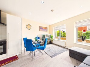 A modern kitchen with white cabinets, a dining area with blue chairs, a TV, and a large window with a view of greenery. - Stylish Watford Stay | Modern | Sleeps 5 (Hertfordshire)