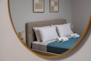 Een net opgemaakt bed met witte lakens en een blauwgroene deken, een bedlampje en twee abstracte kunstwerken aan de muur.