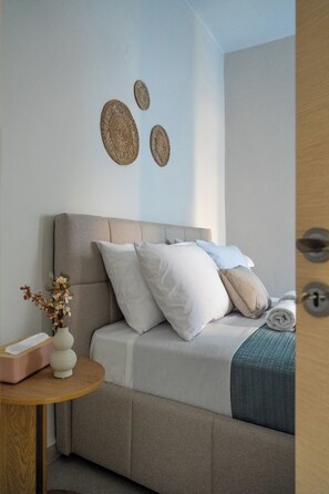 A bedroom with a bed, pillows, a bedside table with a vase, and wall decor. - Elounda Street - Stunning View & Prime location (Schisma Elountas)