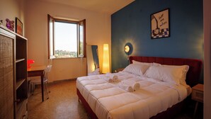 Un lit bien fait, avec du linge de lit blanc, un bureau avec une lampe rouge et une fenêtre donnant sur un beau paysage.