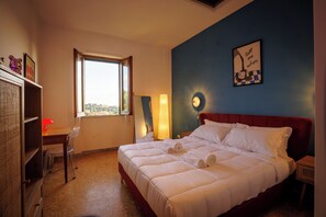 A neatly made bed with white linens, a desk with a red lamp, and a window with a view. - Vrbo Property (Frascati)