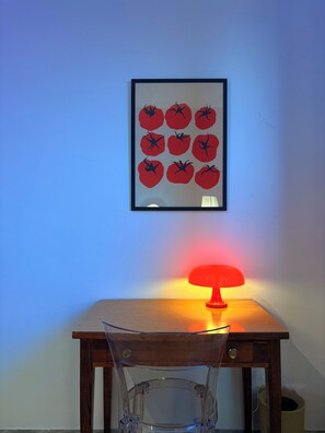 Une œuvre d’art encadrée représentant des tomates, accrochée à un mur, une table avec une lampe rouge et une chaise en verre transparent.