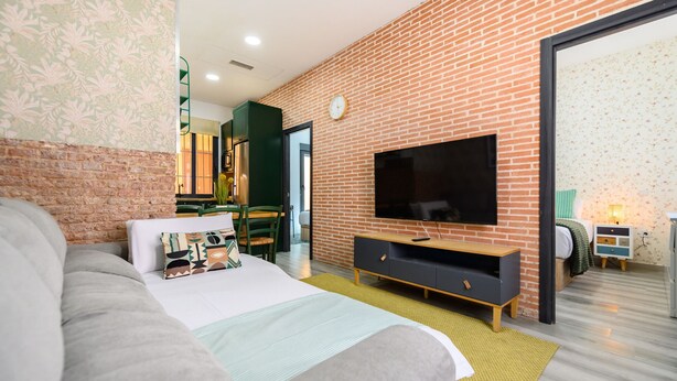 Ein modernes Hotelzimmer mit Backsteinwand, einem großen Bett, einem Flachbildfernseher und einem Essbereich, der durch eine offene Tür sichtbar ist.