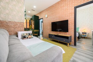 Ein modernes Hotelzimmer mit Backsteinwand, einem großen Bett, einem Flachbildfernseher und einem Essbereich, der durch eine offene Tür sichtbar ist.