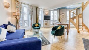 Ein Wohnzimmer mit einer Couch, Sesseln, einem Fernseher, einer Treppe und einem Essbereich.