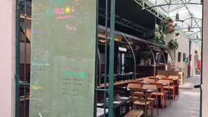 Ein Straßencafé mit einer Kreidetafel, die Speisen und Preise auflistet, Holztische und -bänke sowie hängende Pflanzen.