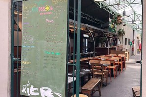 Ein Straßencafé mit einer Kreidetafel, die Speisen und Preise auflistet, Holztische und -bänke sowie hängende Pflanzen.