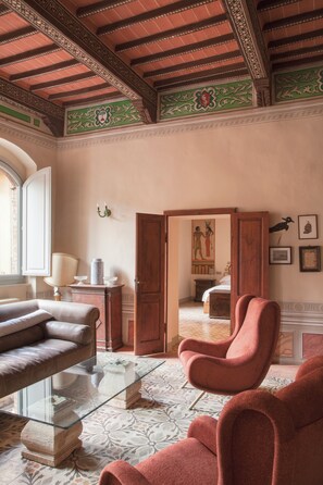 Een kamer met een glazen salontafel, een leren bank, een roze fauteuil en een houten deur die naar een andere kamer leidt.