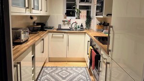 Een kleine keuken met witte kastjes, een houten aanrecht, een inbouwoven en een raam met decoratieve objecten.