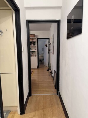 Ein Korridor mit Holzboden, einer weißen Tür links und einem Fenster mit schwarzem Rahmen rechts.