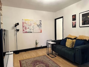 Ein Wohnzimmer mit einem dunkelgrünen Sofa, einem kleinen Beistelltisch, einer Stehlampe, einem großen abstrakten Gemälde und einem Spiegel an der Wand.