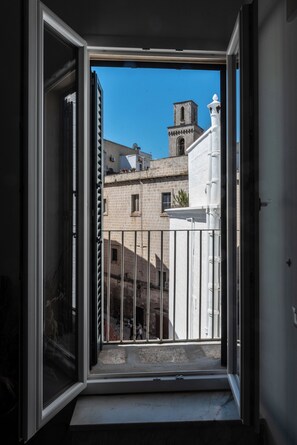 Blick durch ein offenes Fenster auf ein Gebäude mit einem Glockenturm, einem weißen Zaun und einem Balkon.
