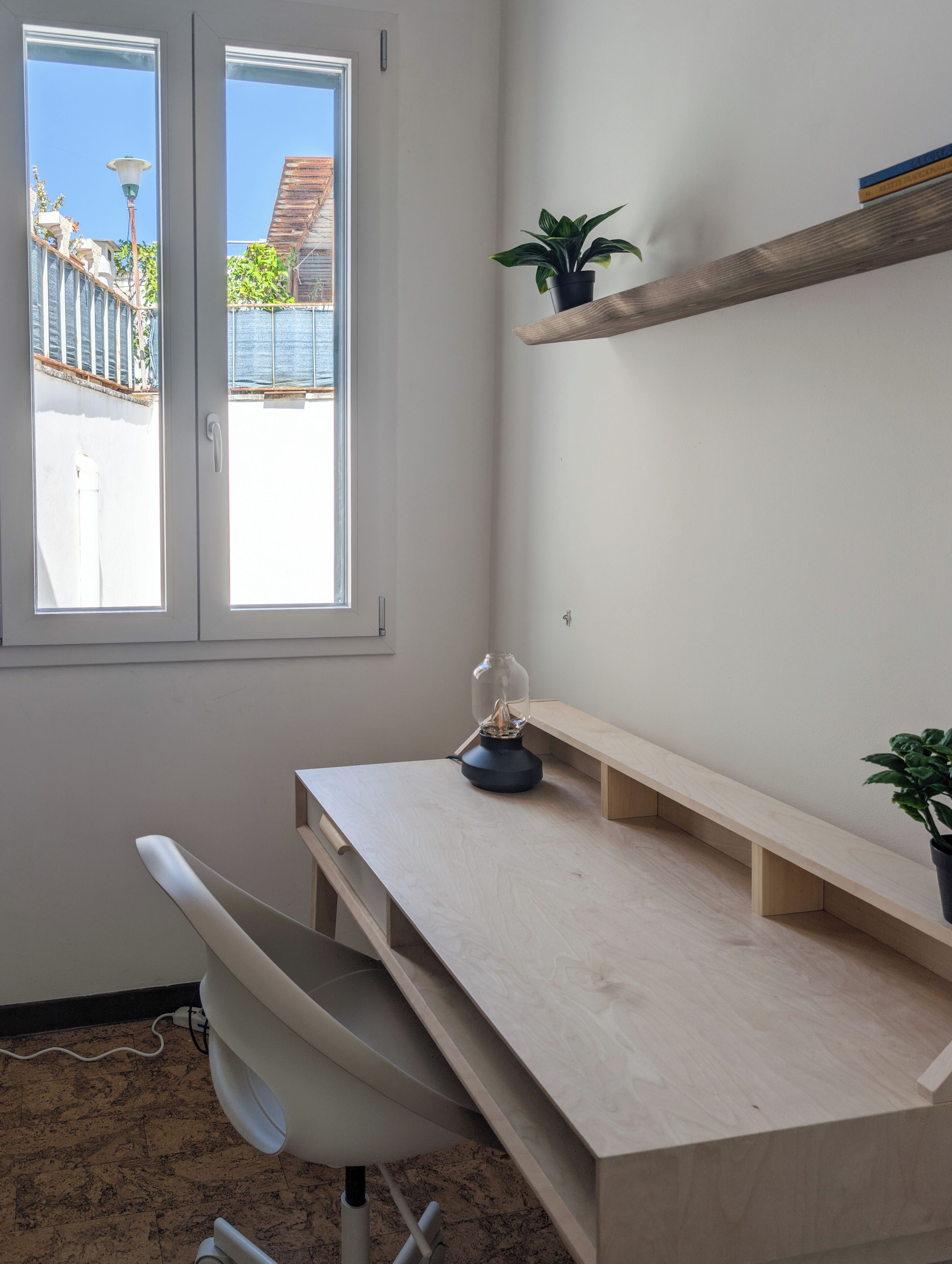 A room with a desk, chair, shelving, and a window with a view of buildings and greenery.