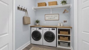 Une buanderie équipée de deux machines à laver à chargement frontal, d’une étagère en bois avec des paniers, d’un panier suspendu, d’une plante en pot et d’un panneau indiquant : « Laver, sécher, plier, répéter ».