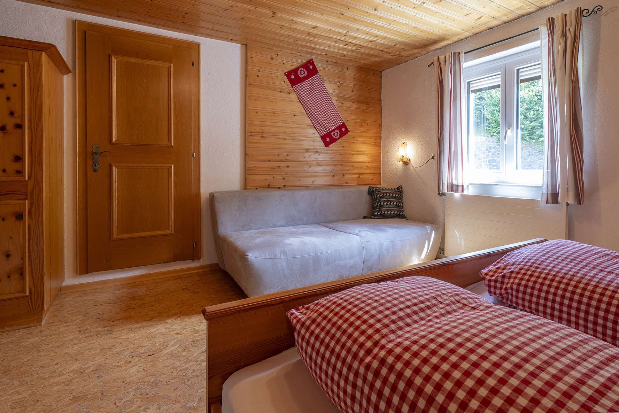 A room with a wooden door, a sofa, a bed with checkered bedding, a wall-mounted light, and a window with curtains.