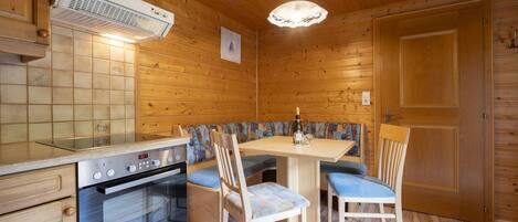 A wooden cabin interior with a kitchen area featuring a stove, a dining table with chairs, and a bench.