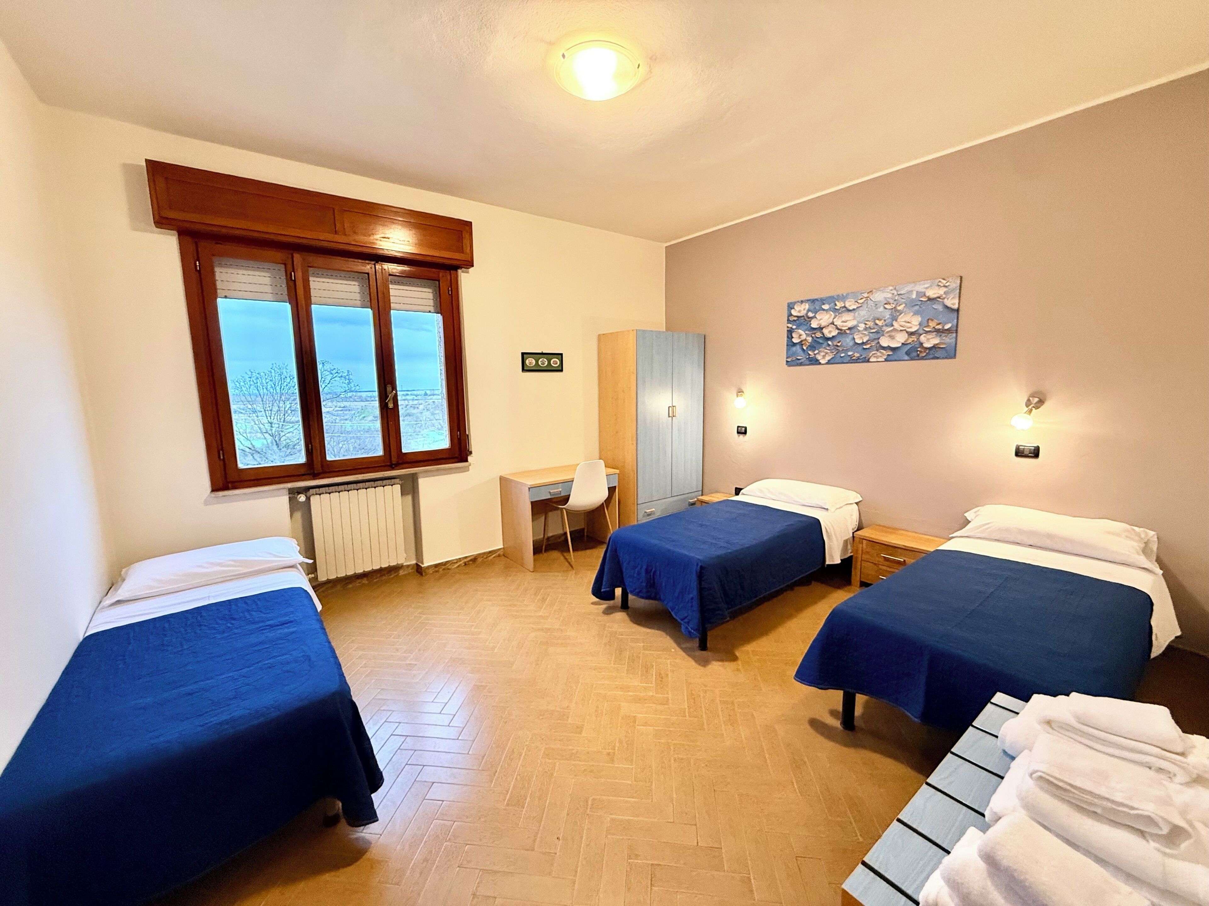 A room with three beds, a desk, a chair, a radiator, and a window with a view.