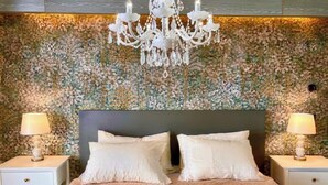 Un dormitorio con papel tapiz floral, un candelabro, dos mesitas de noche con lámparas y una cama con almohadas.