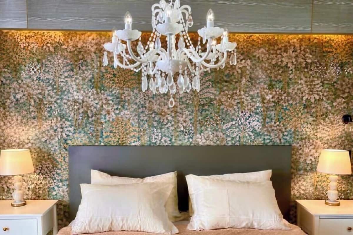Un dormitorio con papel tapiz floral, un candelabro, dos mesitas de noche con lámparas y una cama con almohadas.